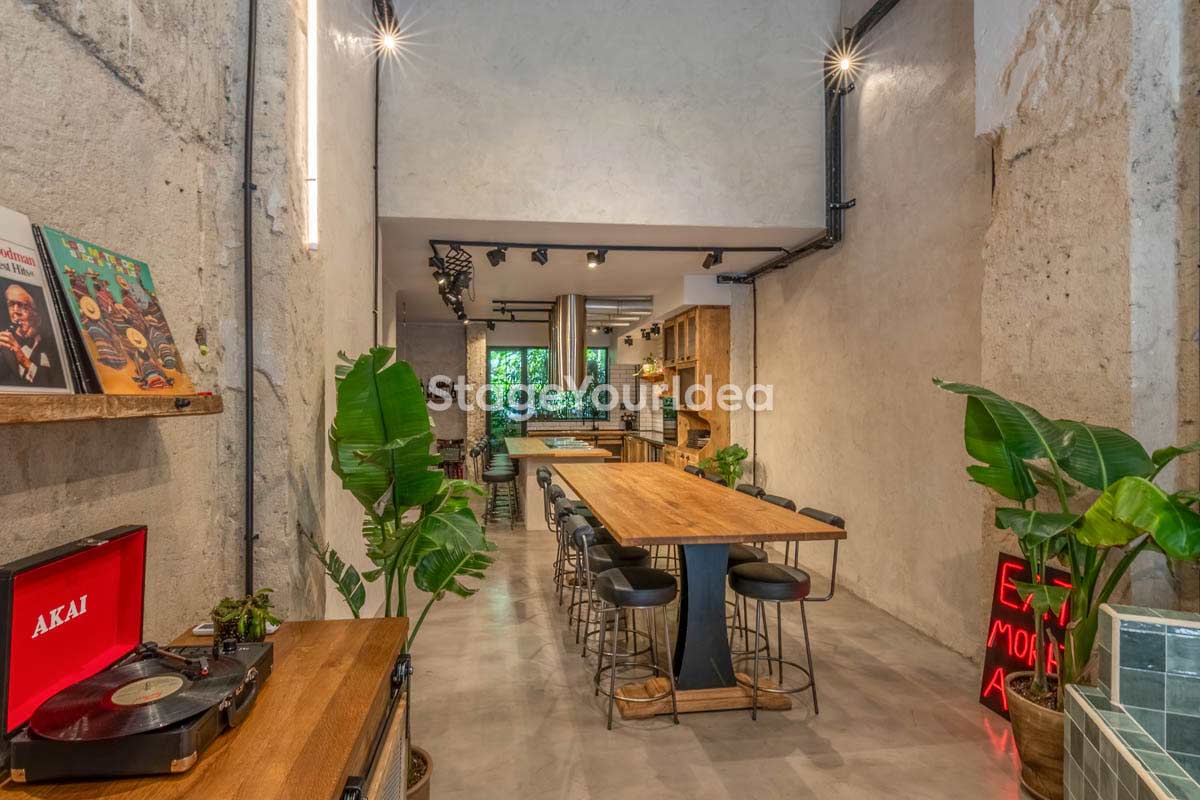 Αστικό events studio στη Θεσσαλονίκη με ξύλινο τραπέζι, ψηλά σκαμπό, φυτά και κουζίνα στο βάθος.